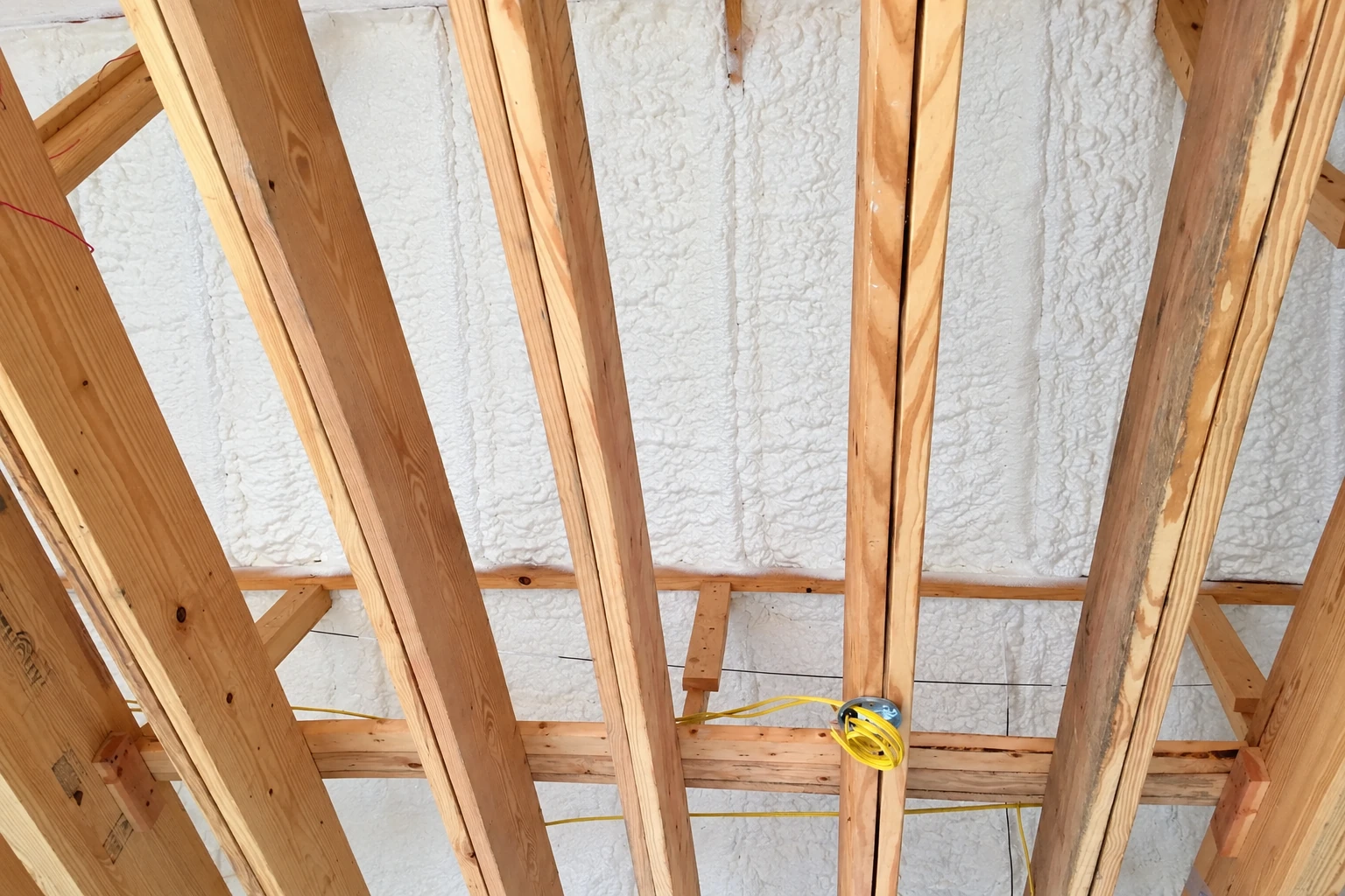View upward between wooden ceiling joists at a surface coated with white spray foam insulation and yellow wiring.