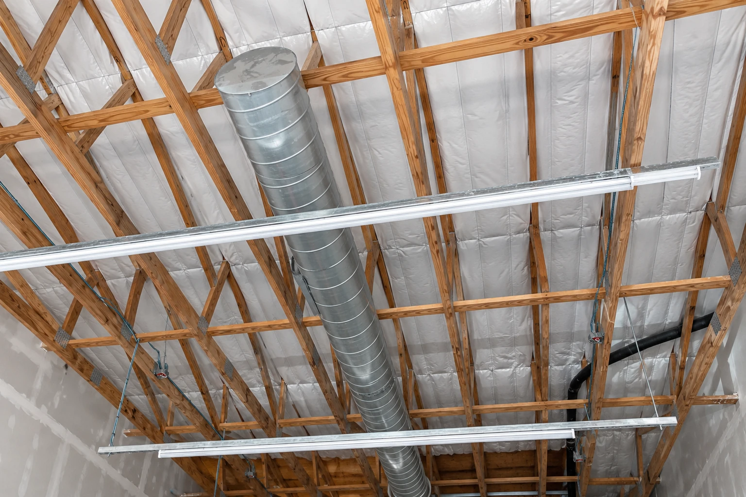 Commercial building ceiling with white insulation between exposed wooden trusses, ductwork, and suspended light fixtures.