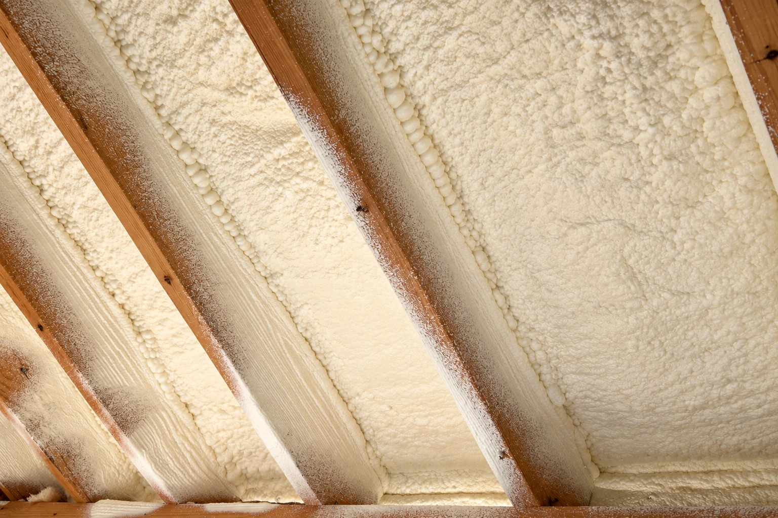Cream-colored spray foam fills the spaces between exposed wooden rafters on a sloped ceiling.