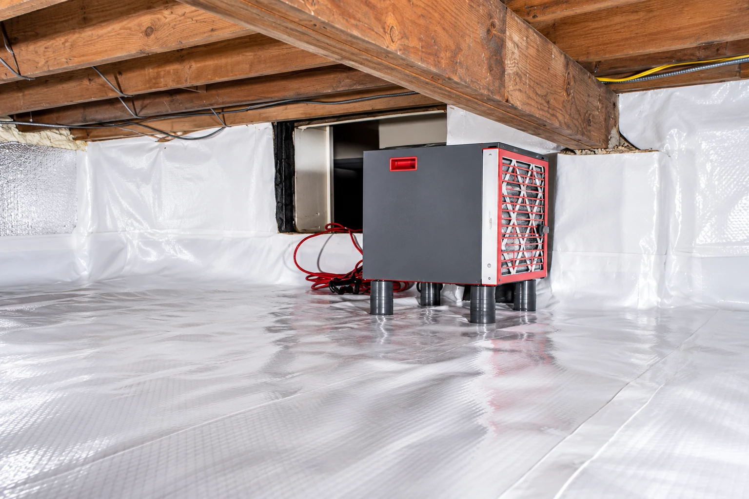 A crawl space has white vapor barrier lining its floor and walls around a gray dehumidifier.